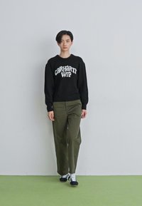 Osoba idąca do przodu, ubrana w czarny sweter Carhartt WIP, oliwkowe spodnie i czarno-białe sneakersy, na tle gładkiej ściany i zielonej podłogi.