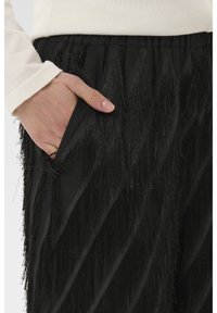 Czarne spodnie z frędzlami wykonane z tkaniny o fakturze, z ukośnymi wzorami i elastycznym paskiem w talii. Dłoń w kieszeni dla kontekstu.