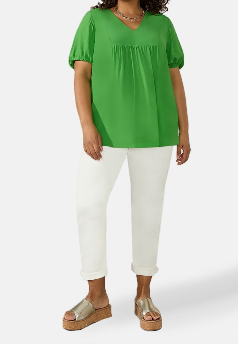 Blusa verde a maniche corte con scollo a V e davanti plissettato, abbinata a pantaloni bianchi con risvolto e sandali metallici con tacco in sughero.