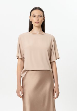 Frau mit glatten dunklen Haaren, die eine beige kurzärmelige Bluse und einen passenden Satinsrock trägt, steht vor einem schlichten weißen Hintergrund.