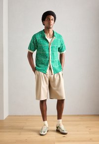 Grønne mønstrede kortærmede skjorte med beige detaljer, parret med beige shorts og sneakers med grønne accenter. Baggrund med lys trægulv.