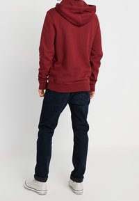 Felpa color bordeaux con cappuccio regolabile, polsini e orlo a coste, abbinata a jeans blu scuro e sneakers bianche. Tessuto morbido, vestibilità casual.