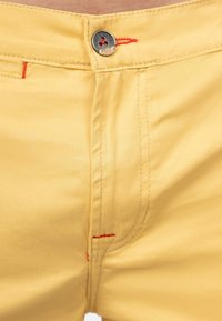 Geel katoenen broeken met een gladde textuur, voorzien van rode stiksels en een metallic knoopaccent. Close-up van het taillebandgebied.