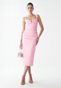 Robe rose ajustée avec un décolleté carré, détail froncé à l'avant et côtés froncés. Le mannequin tient un petit sac à main blanc et porte des sandales à talons transparents.