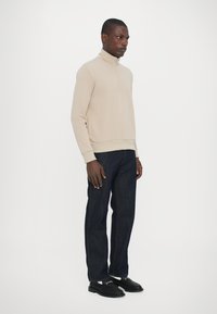 Beige zip-up tröja med hög krage, kombinerad med mörkblå raka jeans och svarta skor, stående mot en vit bakgrund.
