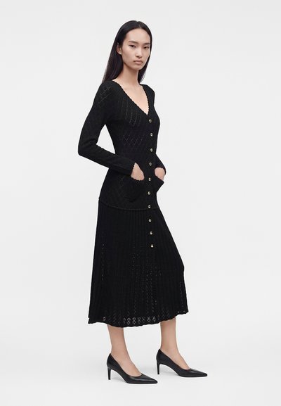 Femme portant une longue robe en maille noire avec des boutons sur le devant et des poches, associée à des chaussures noires à talons hauts pointues.