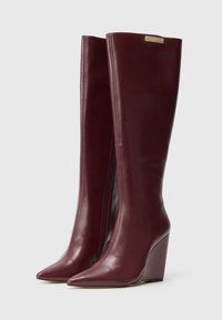 Pair of tall, dark burgundy leather wedge boots with pointed toes and a gold brand tag near the top edge.