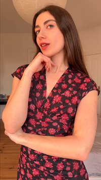 Γυναίκα με μακριά καστανά μαλλιά, φοράει μαύρο φόρεμα με κόκκινο ανθισμένο σχέδιο, ποζάροντας με το χέρι κοντά στο πηγούνι σε ένα καλά φωτισμένο δωμάτιο.