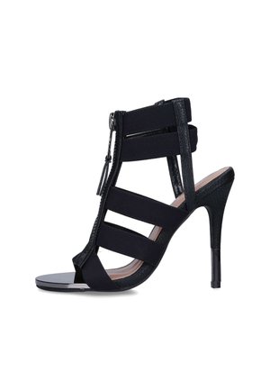Sandale noire à talon haut avec plusieurs brides élastiques, bout ouvert et fermeture éclair à l'avant, vue de profil sur fond blanc.