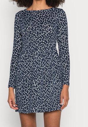 Woman wearing a navy blue dress with long sleeves and white irregular dot pattern, gathered waist, and above-knee length.