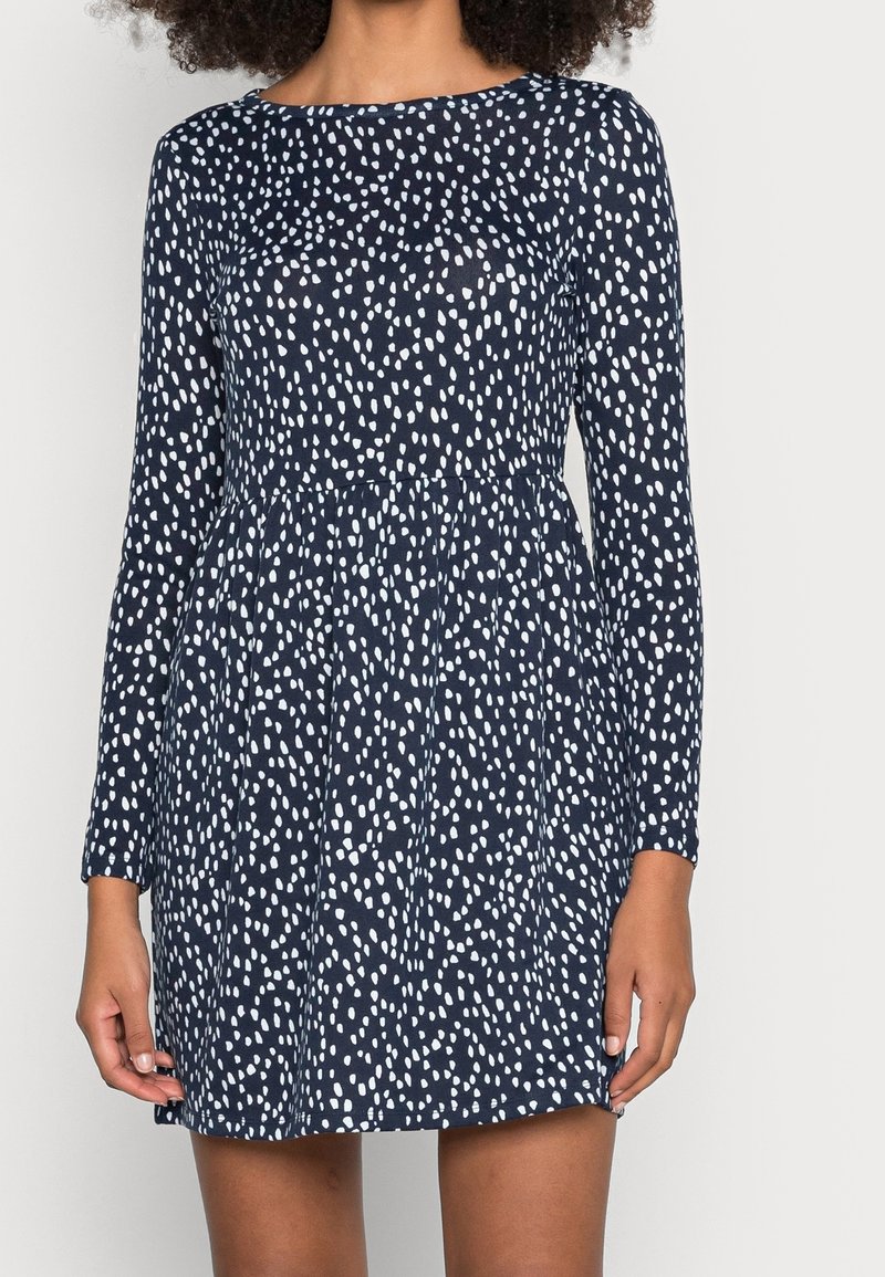Femme portant une robe bleu marine à manches longues avec un motif de points blancs irréguliers, taille froncée, et longueur au-dessus du genou.