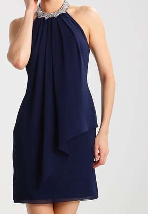 Robe bleu marine sans manches avec encolure licou ornée de strass et détail de jupe asymétrique à couches, portée par une personne debout.