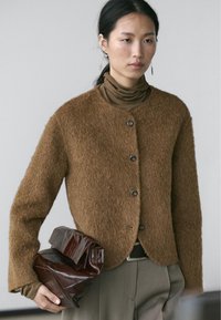 Woman wearing a brown fuzzy cardigan over a brown turtleneck, holding a large dark brown leather clutch bag at her side.