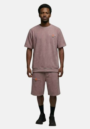 Homme debout portant un ensemble mauve assorti constitué d'une chemise à manches courtes et d'un short avec des poches zippées, des chaussettes noires et des baskets noires.
