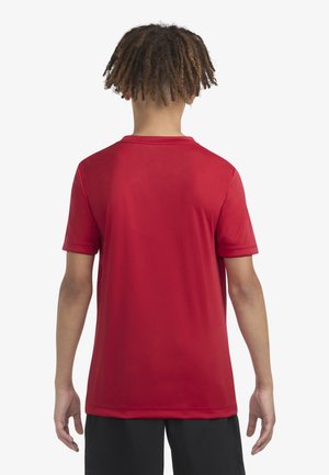 Person mit kurzen lockigen Haaren steht mit dem Rücken zur Kamera, trägt ein schlichtes rotes Kurzarmshirt und schwarze Shorts vor weißem Hintergrund.