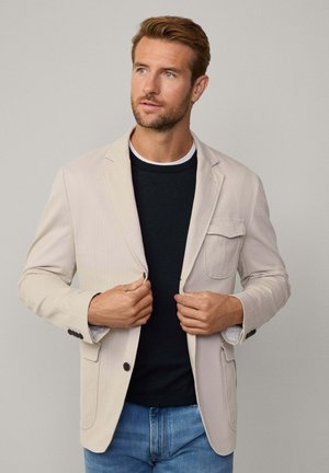 Ljusbeige kavaj med en enda bröstficka, notch-lapel, och knappmanschetter, bärs över en mörk marinblå crewneck-tröja och blå jeans.