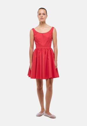 Femme debout portant une robe rouge sans manches longueur genou avec un corsage ajusté et une jupe évasée, associée à des chaussures légères à enfiler de couleur claire.