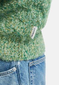 Pull en maille vert texturé avec une étiquette blanche GANT glissée dans la poche d'un jean en denim bleu.