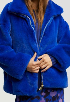 Persona que lleva una chaqueta vibrante de piel sintética azul con una cremallera dorada parcialmente abierta sobre una falda o vestido morado con estampado.