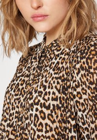 Blusa animal print in beige e nero con dettagli arancioni. Presenta un collo, tessuto plissettato e chiusura con bottoni, creando un aspetto tessurizzato.