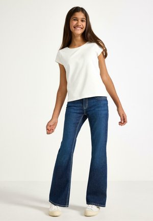 Vit bomullst-shirt med korta ärmar kombinerad med blå utsvängda jeans. Jeansen har synlig sömnad och är lätt slitna vid fållen.