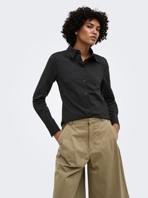 Femme aux cheveux bouclés portant une chemise noire boutonnée et un pantalon large beige taille haute, les mains dans les poches, sur un fond clair.