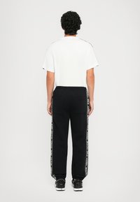Homem com cabelo encaracolado escuro, vestindo uma t-shirt branca, calças de treino pretas com listras laterais em texto e sapatilhas pretas, de pé, de costas, num fundo branco.