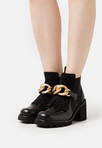 Černé kožené kotníkové boty s robustním podpatkem, zdobené zlatým řetízkem a sukněným třásněmi. Hladký povrch s texturovanými prvky.