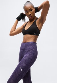 Femme en forme ajustant son élastique à cheveux, portant un soutien-gorge de sport noir et des leggings violets à motifs avec des logos Puma, sur un fond clair uni.