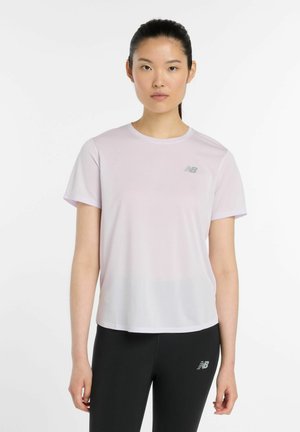 Jonge vrouw met zwart haar vastgebonden, draagt een lichtroze shirt met korte mouwen en zwarte leggings met New Balance-logo's op een witte achtergrond.