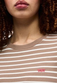 Κοντινό πλάνο ενός ατόμου που φοράει πουκάμισο με μπεζ και λευκές ρίγες και κόκκινο λογότυπο Levi's στο στήθος.