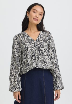 Femme portant un chemisier à manches longues à motif floral noir et blanc et une jupe taille haute bleu foncé devant un fond clair uni.