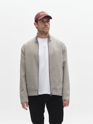 Light gray jacket with a high collar and front zipper, worn over a white shirt. Paired with black pants and a maroon cap with white text.