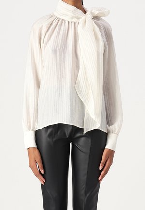 Femme portant une blouse blanche transparente à manches longues avec un grand nœud au cou, associée à un pantalon en cuir noir.