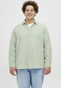 Camisa de manga larga verde claro con botones en el frente y cuello, llevada sobre una camiseta blanca; combinada con jeans de mezclilla azul claro. Corte y textura casual.