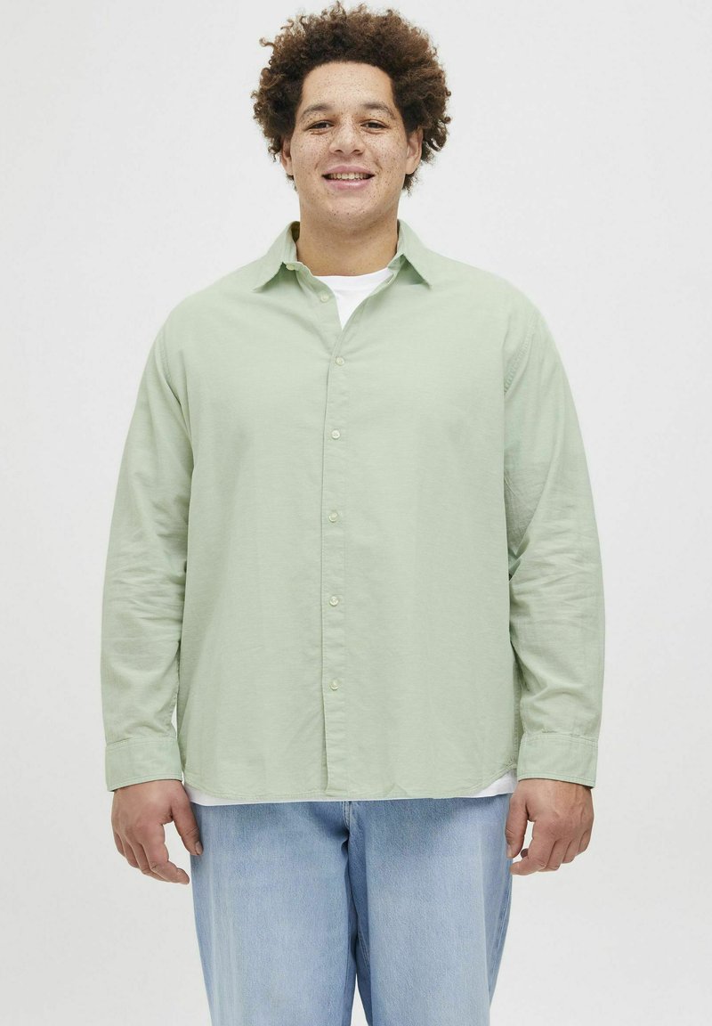Camisa de manga larga verde claro con botones en el frente y cuello, llevada sobre una camiseta blanca; combinada con jeans de mezclilla azul claro. Corte y textura casual.