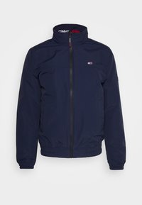 Granatowa wiatrówka z wysokim kołnierzem, elastycznymi mankietami i dole; posiada czarny zamek błyskawiczny oraz małe logo na piersi.