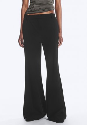 Persoon die zwarte broek met hoge taille en wijde pijpen draagt en een donkergrijze mouwloze top die is ingestopt, staand tegen een effen achtergrond.
