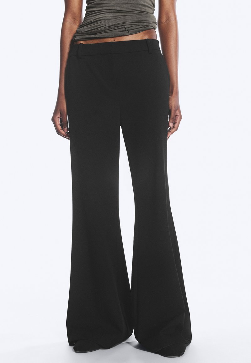Personne portant un pantalon noir taille haute à jambes larges et un haut sans manches gris foncé rentré, debout devant un fond uni.