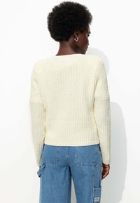 Un pull en tricot couleur crème avec un motif texturé, un ourlet côtelé et des manches longues, associé à un jean bleu ample.