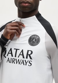 Άνδρας που φοράει μαύρο και λευκό αθλητικό μπλουζάκι του Παρί Σεν Ζερμέν με φερμουάρ και το λογότυπο «Qatar Airways», ρυθμίζοντας το γιακά.