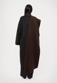 Abrigo largo y de gran tamaño con un diseño dividido que combina telas negra y marrón oscuro, cuello alto y materiales texturizados.