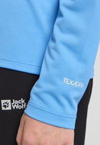 Lichtblauwe langemouwen shirt met een textuurstof, voorzien van het witte "TEXADRI" logo op de mouw. Zwarte broek met het zichtbare witte "Jack Wolfskin" logo.