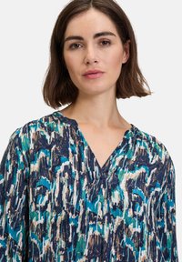 Blouse à motifs avec un décolleté en V et des manches longues. Le tissu présente un mélange de tons bleus, verts et neutres avec un design texturé.