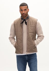 Quiltad beige väst med dragkedja framtill, sidofickor och en texturerad, ribbad krämfärgad tröja under, tillsammans med blå jeans och en svart halsduk.