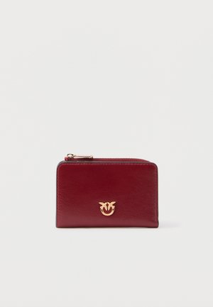 Piros bőr pénztárca arany színű logóemblémával az elején, egyszínű világosszürke háttér előtt.