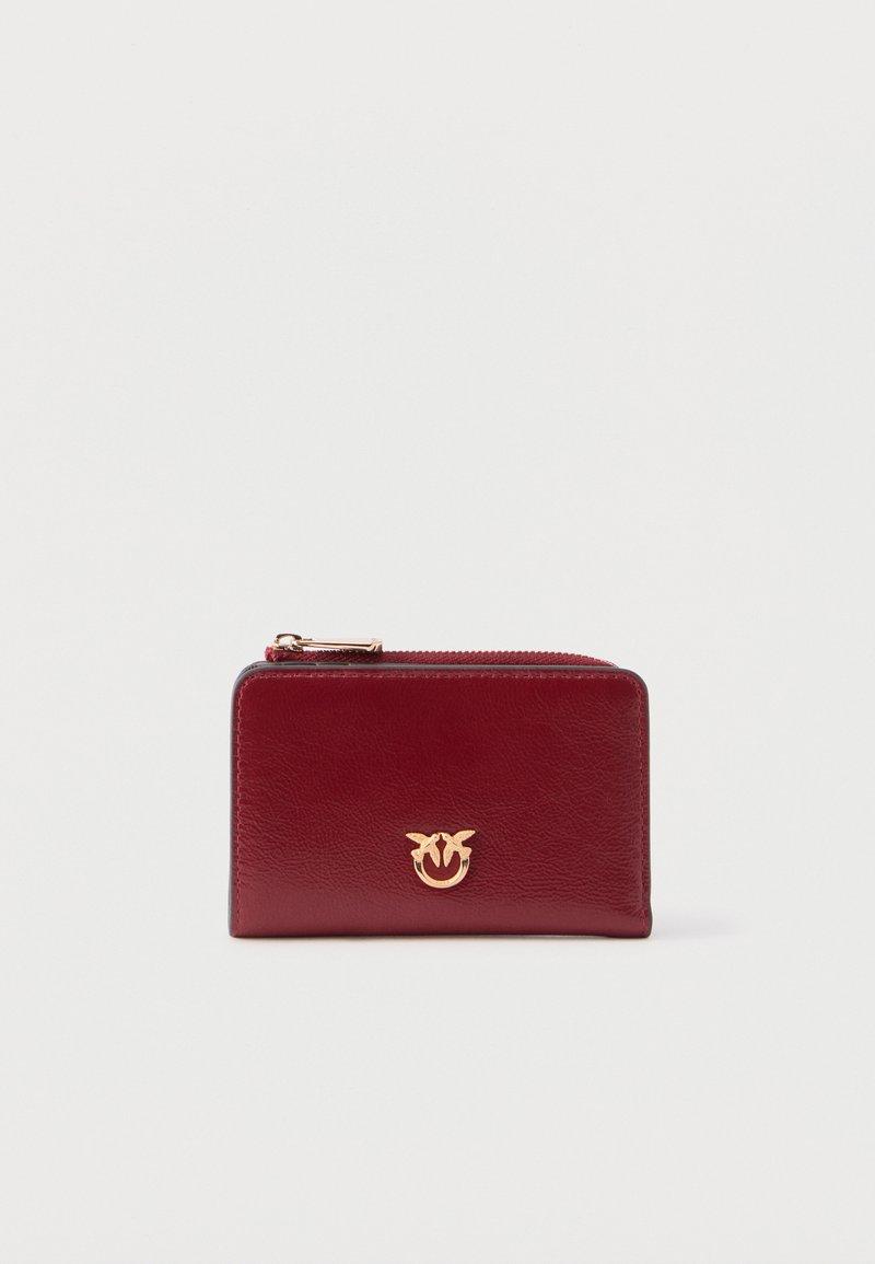 Κόκκινο δερμάτινο πορτοφόλι με φερμουάρ και χρυσόλογο με έμβλημα στο μπροστινό μέρος, τοποθετημένο σε απλό ανοιχτό γκρι φόντο.