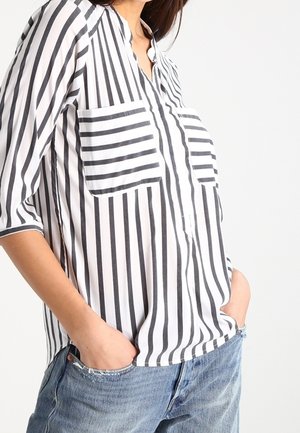 Femme portant une chemise blanche ample avec des rayures noires verticales et horizontales, les mains dans les poches d'un jean en denim bleu sur un fond clair.