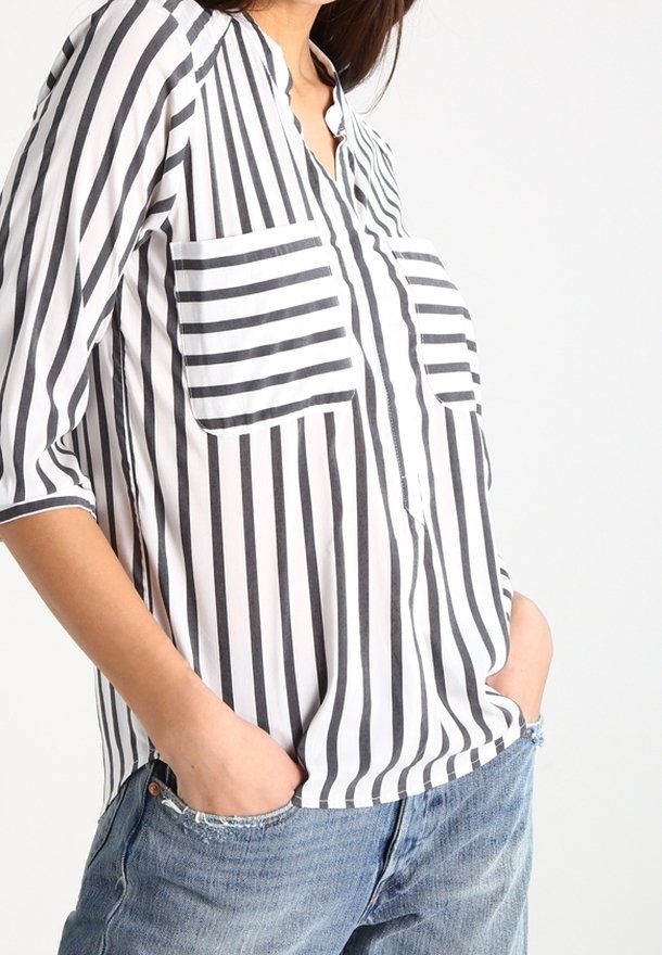 Femme portant une chemise blanche ample avec des rayures noires verticales et horizontales, les mains dans les poches d'un jean en denim bleu sur un fond clair.