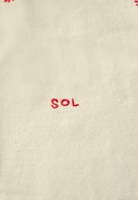 Krémová tkanina s jemnou diagonálnou textúrou má červené vyšívané písmená tvorené slovom "SOL," umiestnené centrálně.